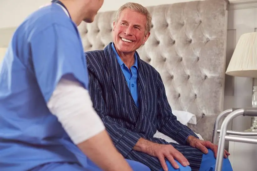 Auf dem Bild ist ein älterer Herr zu sehen, welchem geholfen wird, aus dem Bett aufzustehen.